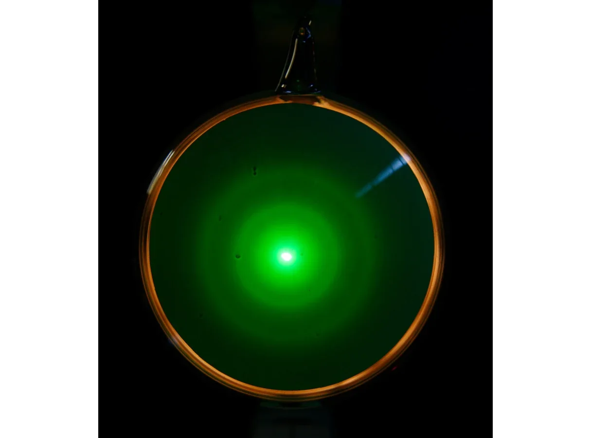 Photograph of the glowing green screen of an electron diffraction tube in a dark room. A bright central spot is surrounded by faint concentric rings.
