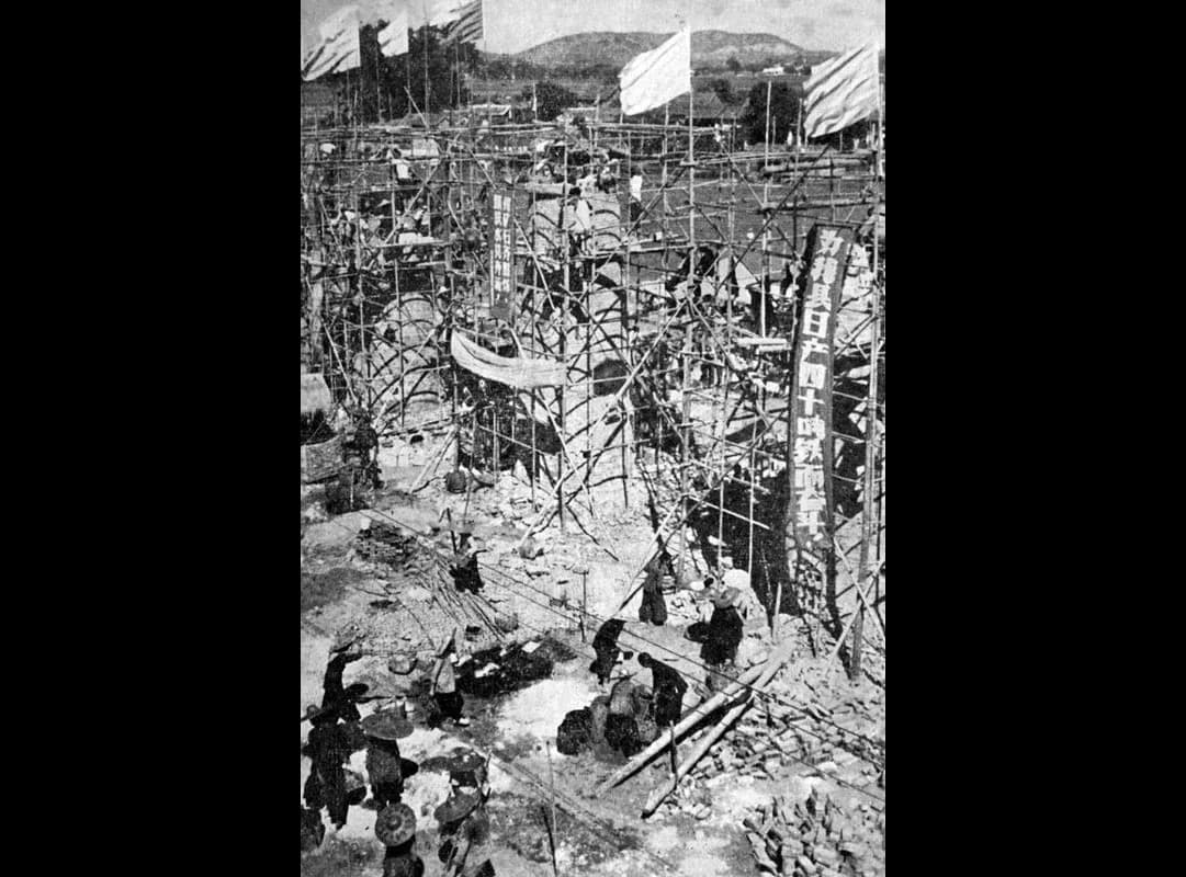 Black-and-white photograph of a large outdoor site during the Great Leap Forward: brick furnaces being built inside bamboo scaffolding under flags and a tall slogan banner, with workers in hats carrying bricks and materials in the foreground.