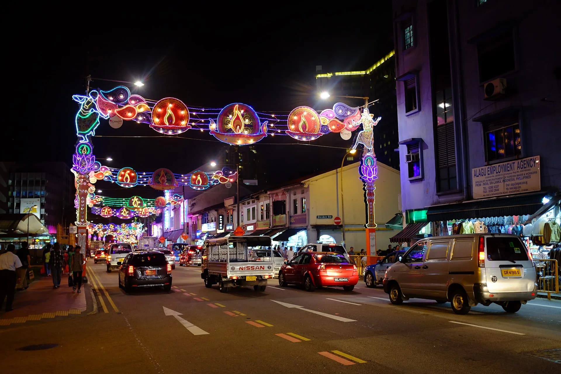 Serangoon Road in Little India, Singapore, during Deepavali 2013: illuminated arches with oil lamps, peacocks and dancers over a street of low shophouses, a clothing shop and an employment agency for workers from India and Sri Lanka.