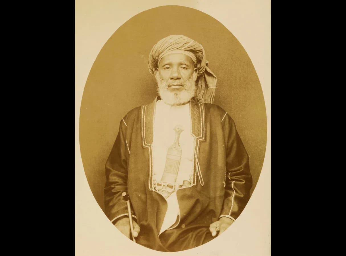 Sepia oval studio photograph of Tippu Tip, an older man with a white beard and a turban, seated in a dark embroidered robe over a white tunic, with a curved dagger at his belt and a cane in his hand.
