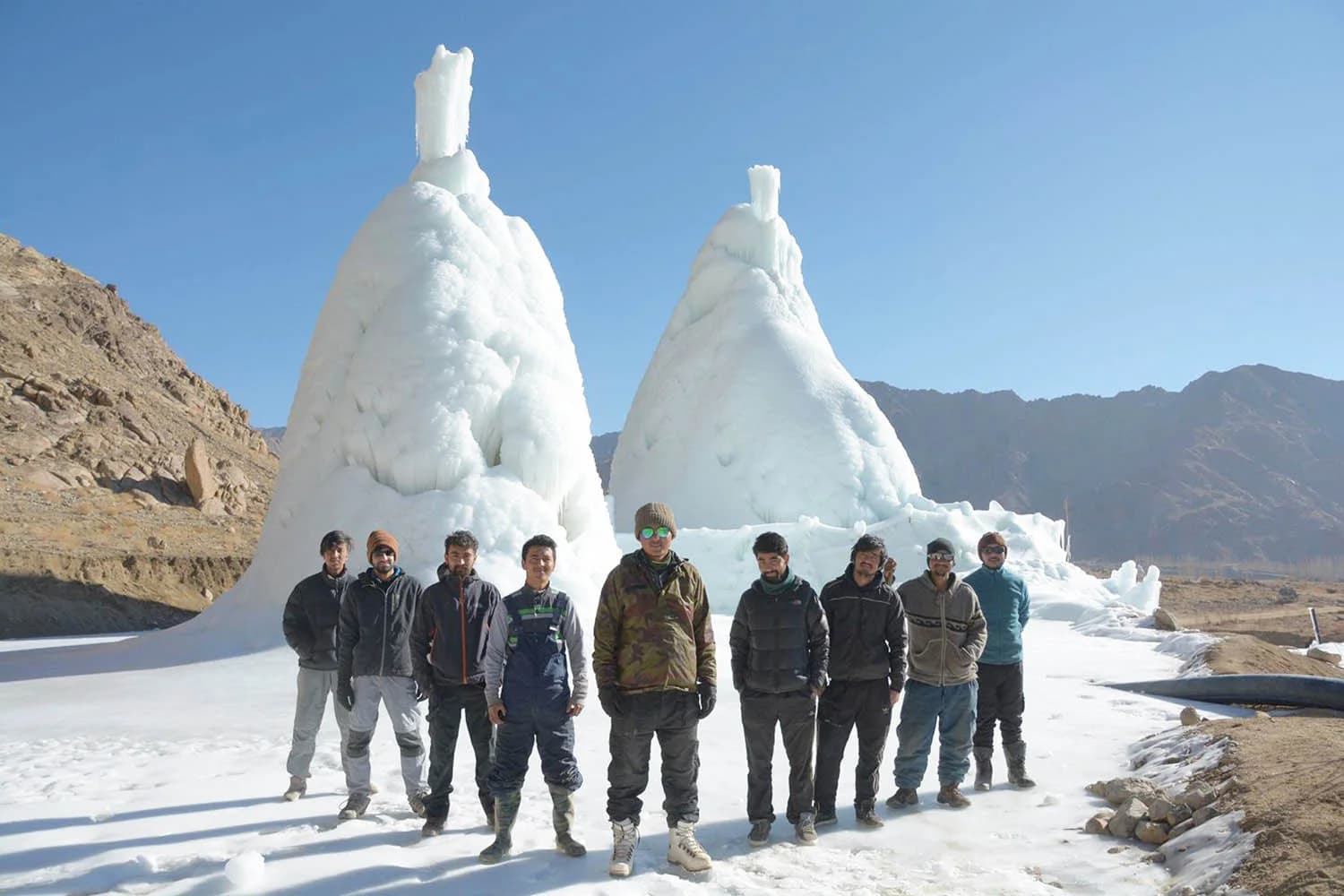 Two tall cones of ice, ice stupas, several times the height of the group of people standing in front of them, on a frozen field in a dry mountain valley in Ladakh, with a water pipe at the right.