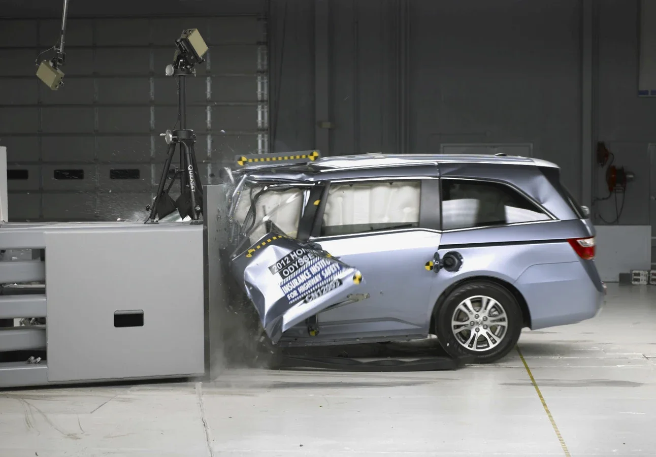 A minivan hitting the corner of a rigid barrier in a crash test. Its front corner is crushed and folded back, and the side curtain airbags have inflated inside the windows.