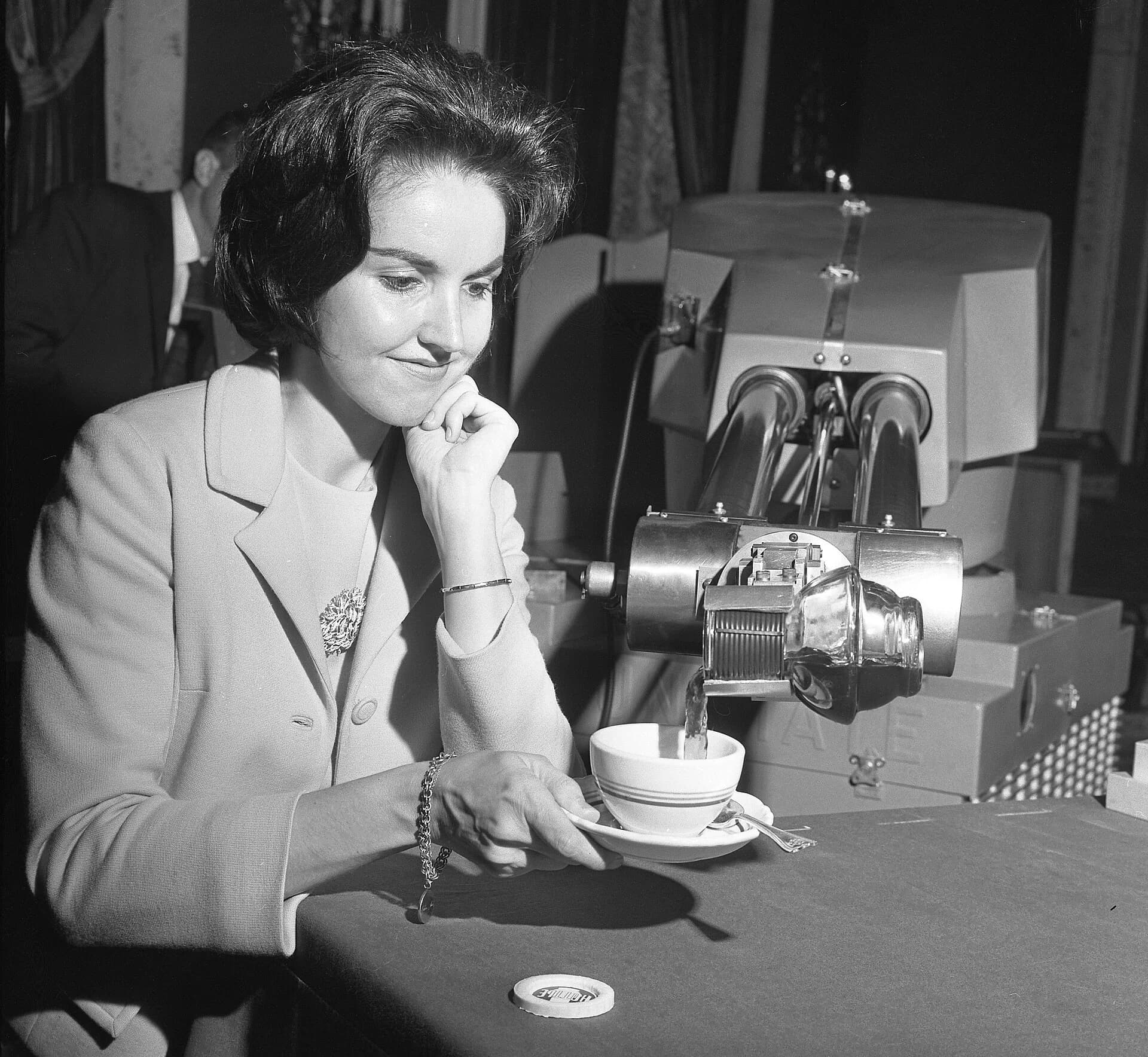 Black-and-white 1967 photograph of a large Unimate robot arm pouring coffee into a cup held by a seated woman at the Biltmore Hotel, Los Angeles.
