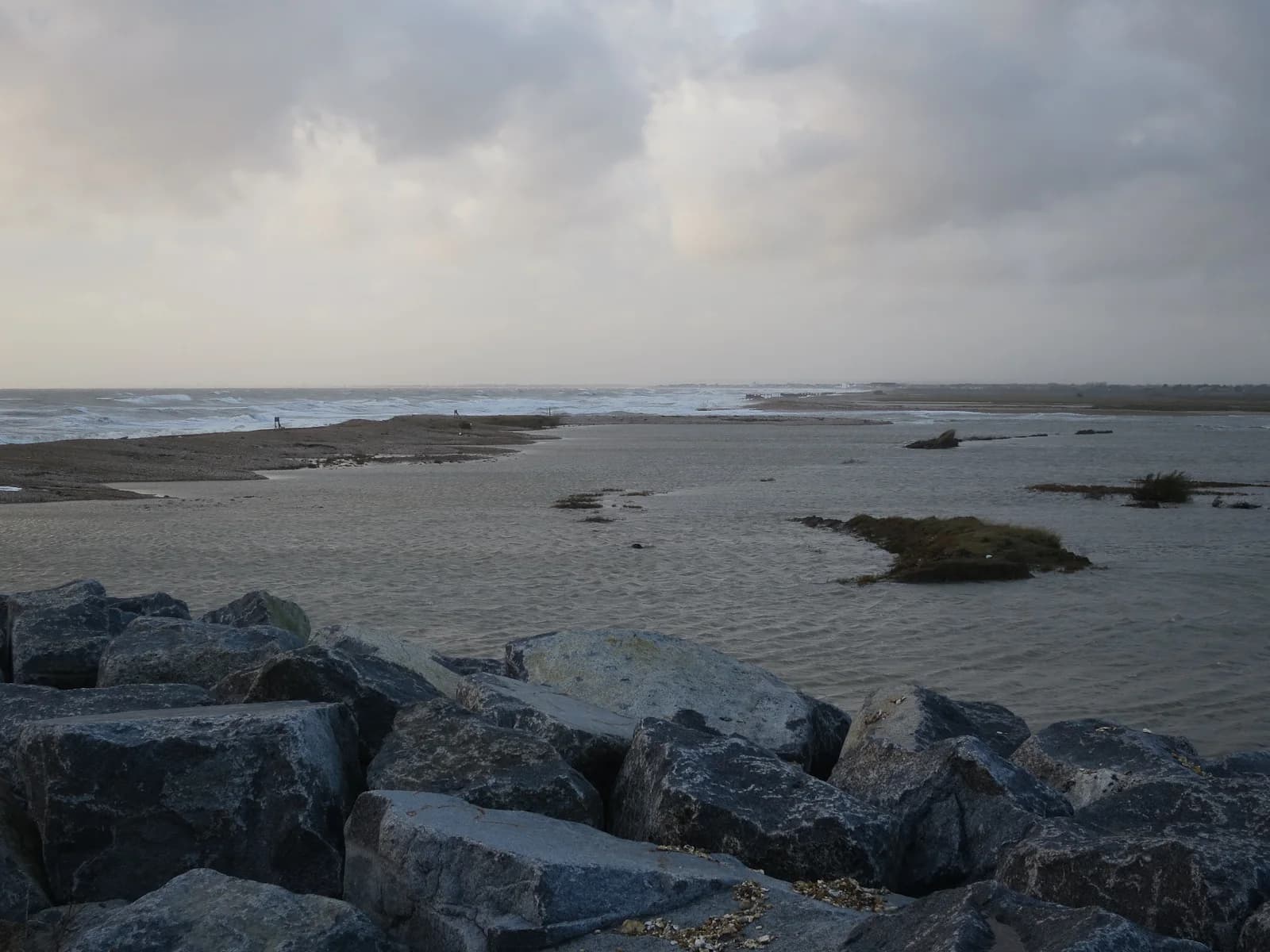 Photograph of the Medmerry managed realignment site, West Sussex: rock armour in the foreground, a breached shingle bank and the open sea behind, and former low-lying farmland now flooded by the tide with patches of vegetation.
