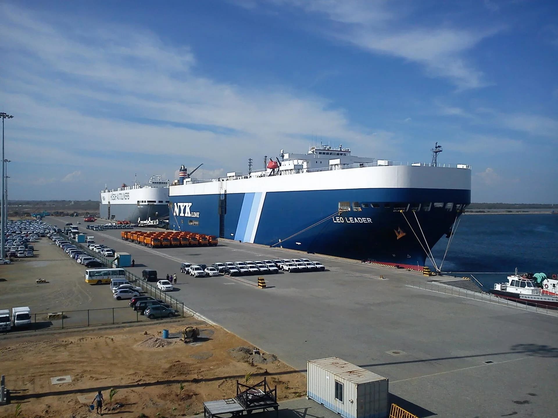 Photograph of Hambantota Port, Sri Lanka: two large car-carrier ships moored at a long concrete quay, with rows of newly unloaded cars and trucks parked on the dockside and open sea to the right.