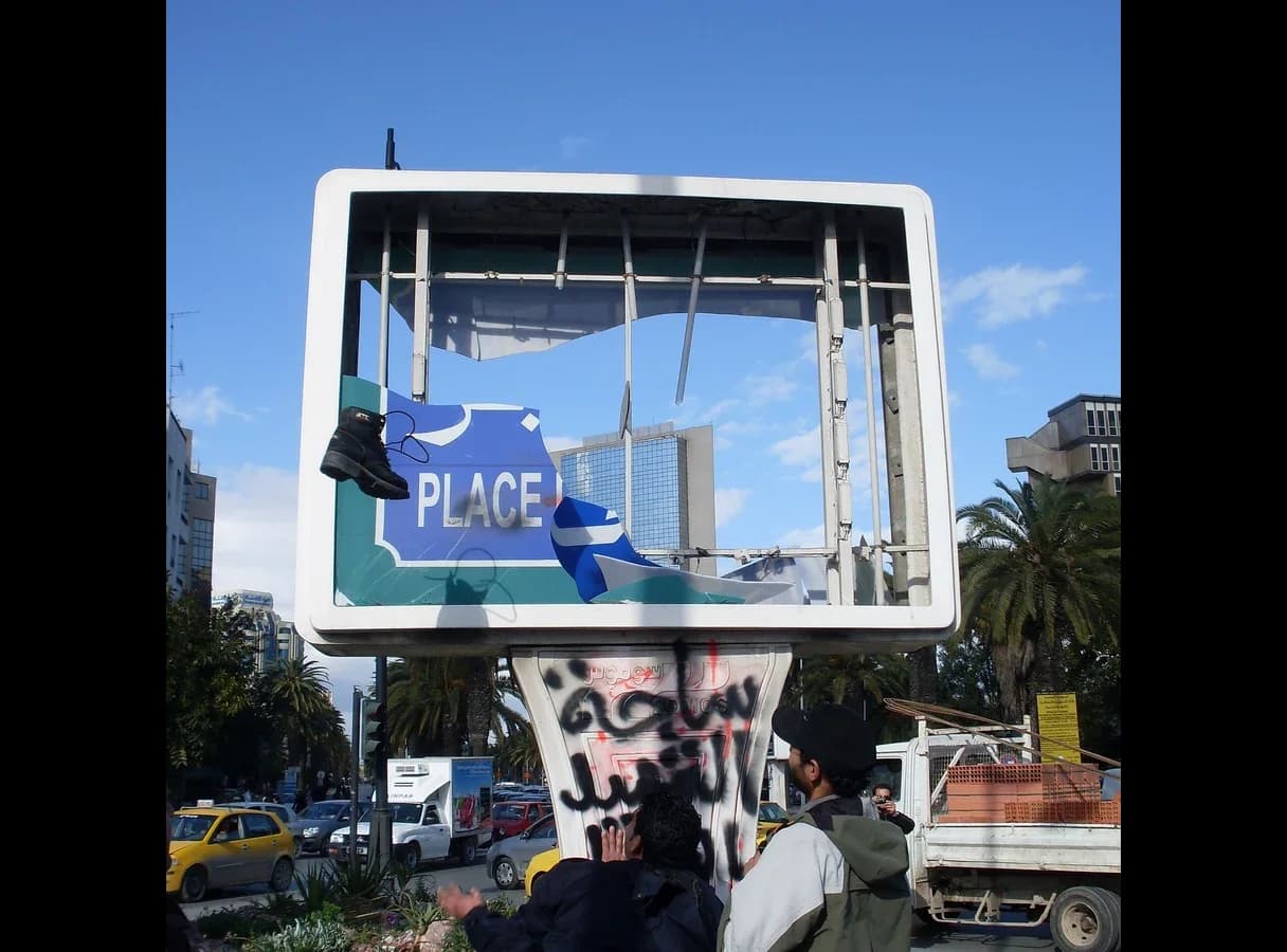 Photograph of a large roadside sign frame in Tunis whose name panel has been torn away, leaving only the word PLACE; a boot hangs from it, the base is covered in graffiti, and a tall glass tower stands behind.