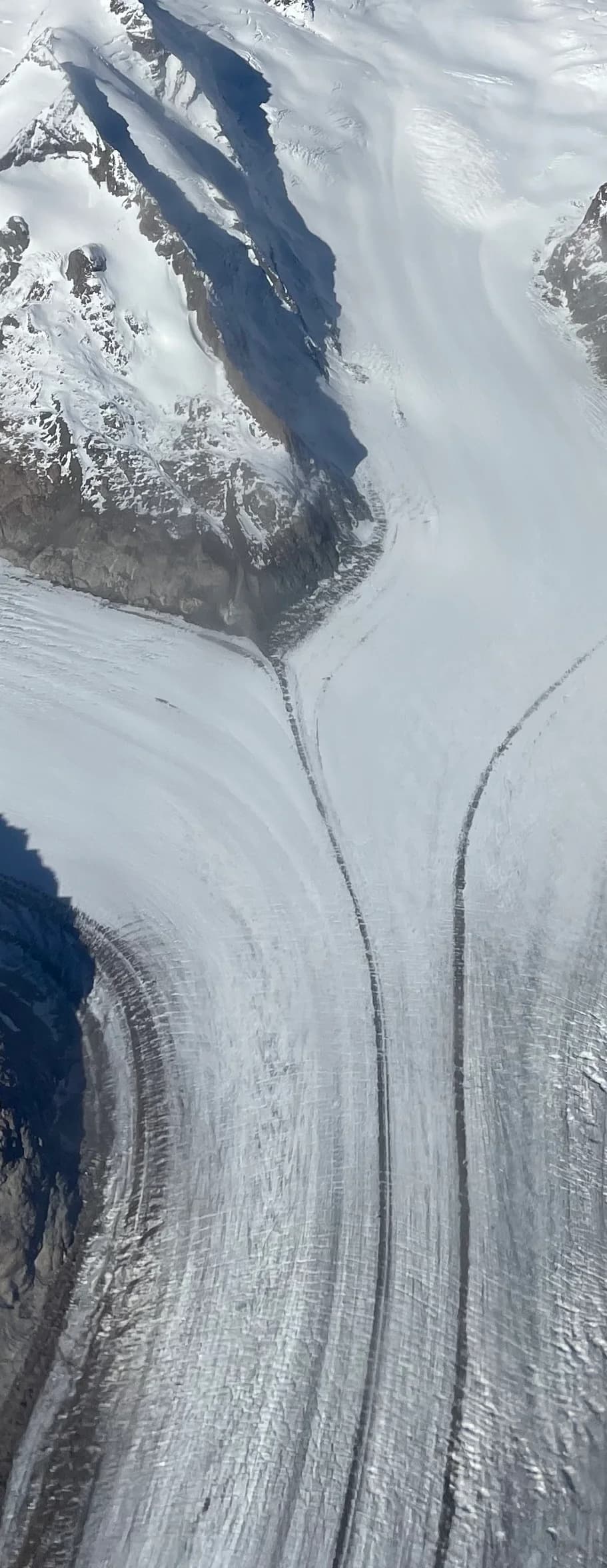 Aerial view of the Great Aletsch Glacier below Konkordiaplatz: tributary ice streams merge, and two thin dark bands of rock debris, medial moraines, run down the white glacier surface parallel to its flow, with a dark lateral moraine along the edge.