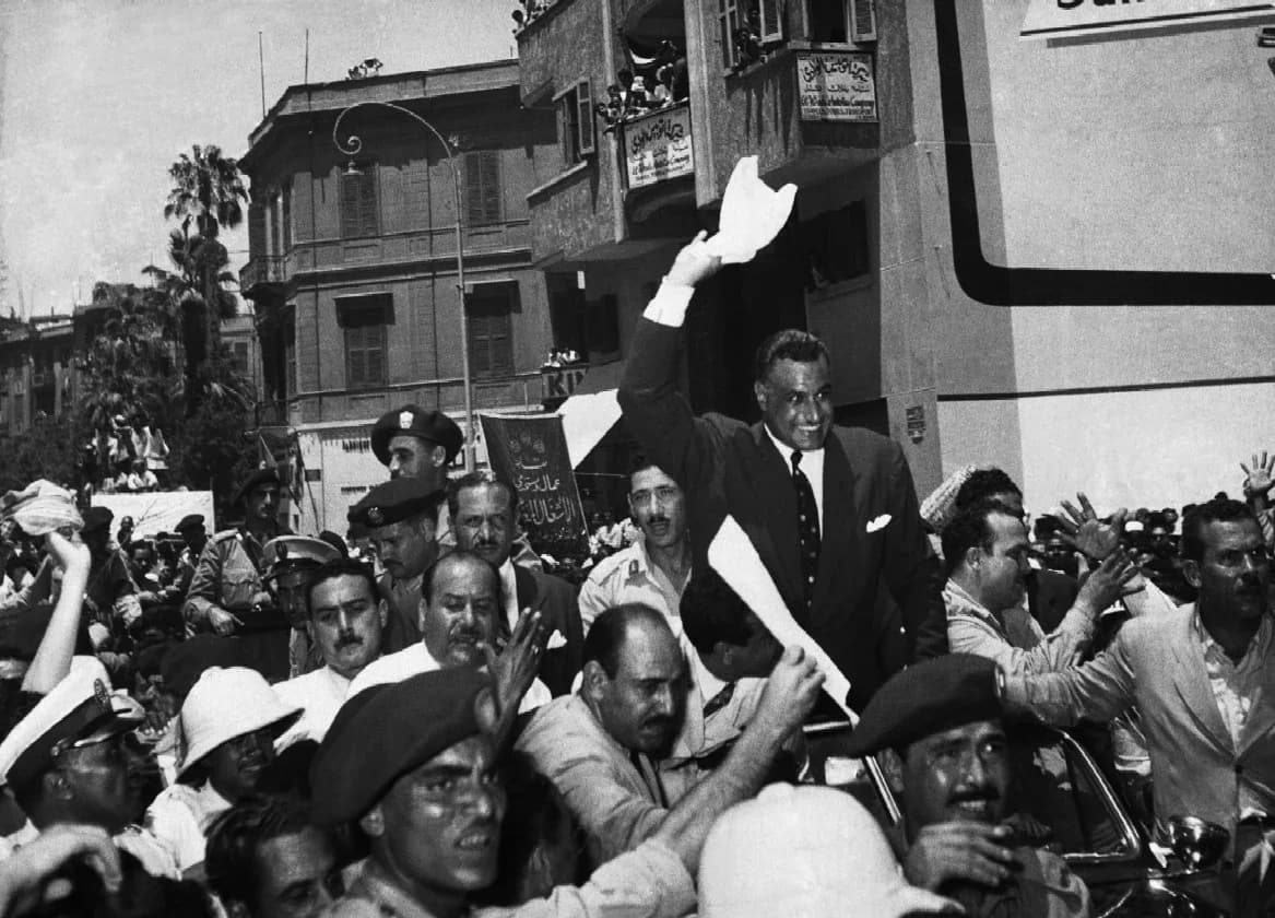 Black-and-white photograph of Gamal Abdel Nasser standing in an open car and waving, surrounded by a dense crowd of men, police and officers who reach towards him and cheer.