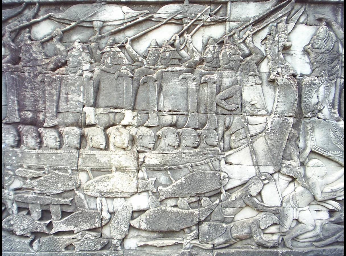 Stone bas-relief showing a long war boat full of armed soldiers with distinctive headdresses, with bodies and fish in the water below.