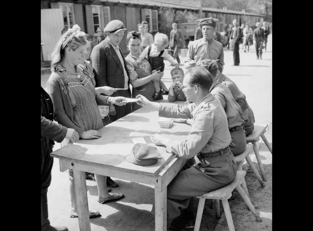 Black-and-white photograph of a Polish family, a young woman, a man, a woman holding a small child and two children, standing at a wooden table while British soldiers seated on stools hand them papers. Wooden barrack huts stand behind them.