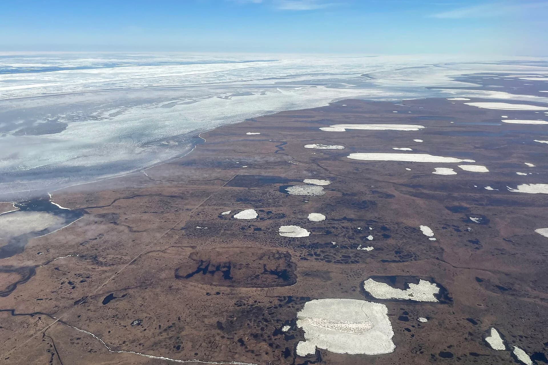 Aerial view of flat, treeless Arctic tundra on the coast of northern Alaska: brown low vegetation dotted with thaw lakes, some still ice-covered, beside sea ice.