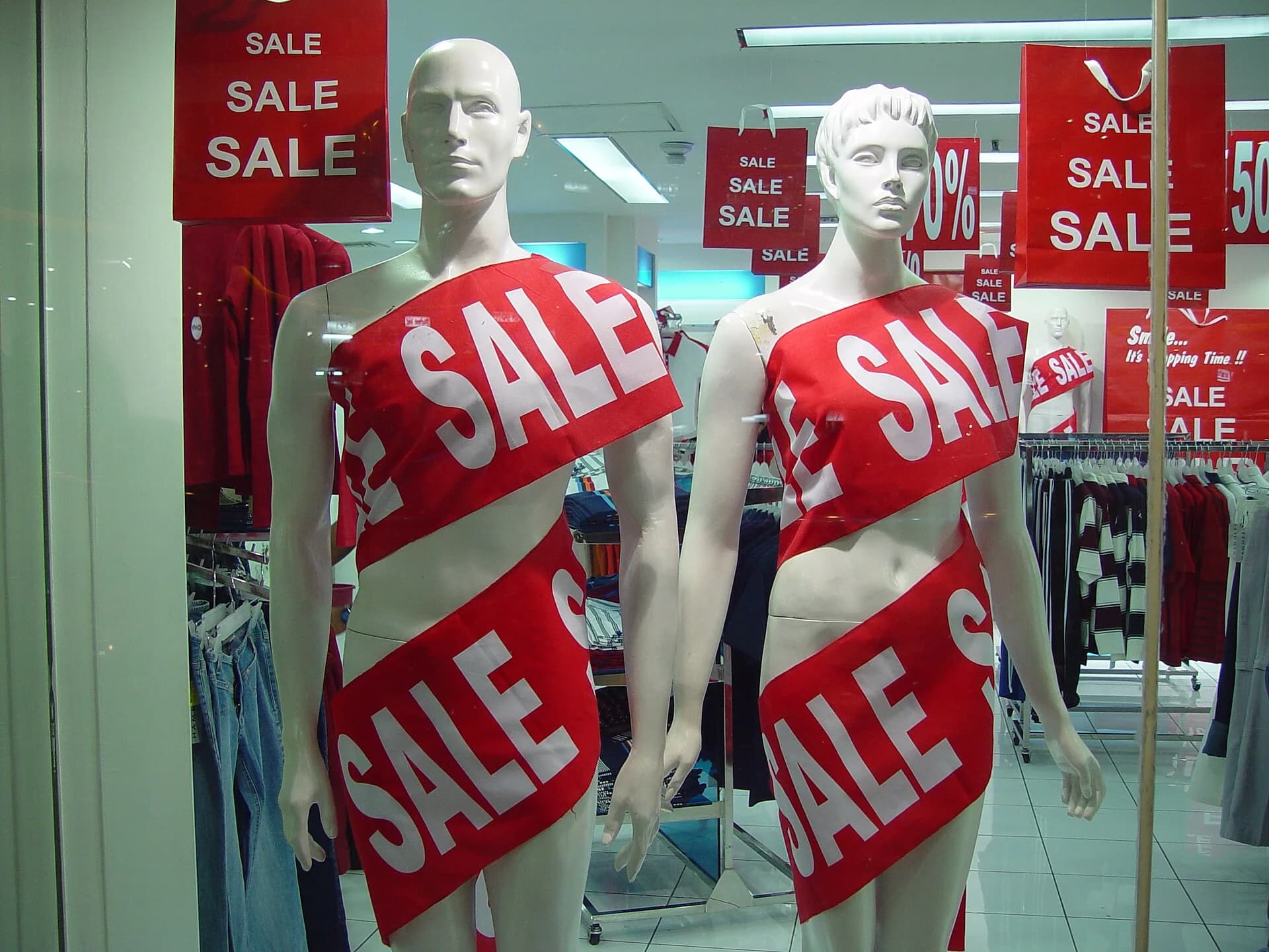 Two shop-window mannequins wrapped in red SALE banners, with more SALE and percentage-off signs hanging in the clothing shop behind them, in a Jakarta shopping mall.