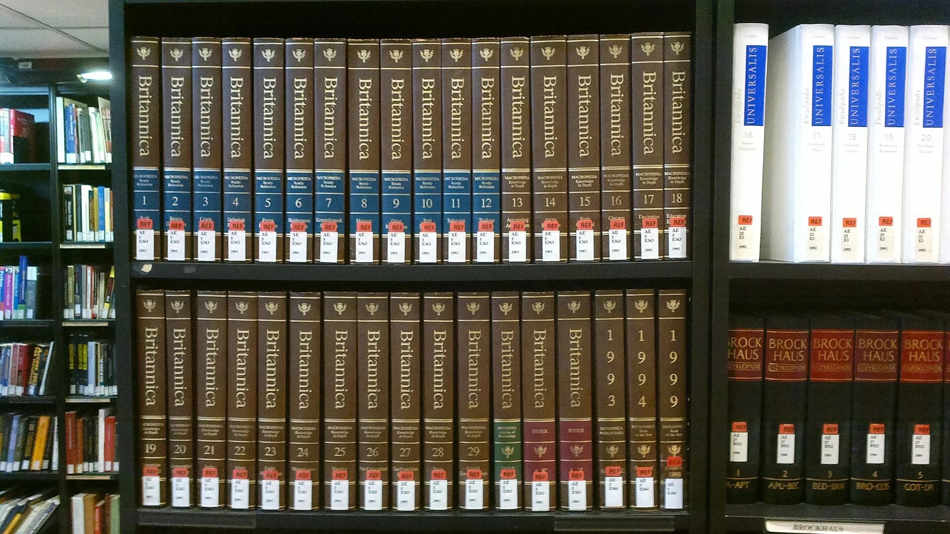 Two library shelves holding the brown bound volumes of the 15th edition of the Encyclopaedia Britannica (1993 printing), numbered 1 to 29 plus index and yearbook volumes, beside sets of other printed encyclopaedias.
