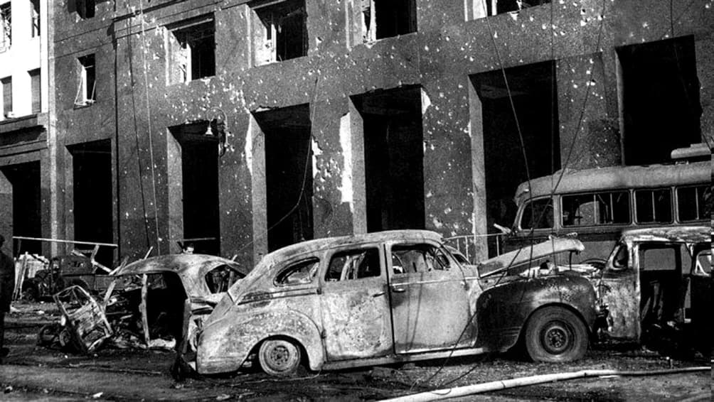 Black-and-white photograph of burnt-out cars and a damaged bus in front of a stone building in Buenos Aires whose walls and window openings are pitted by bullets and shrapnel, after the bombing of 16 June 1955.