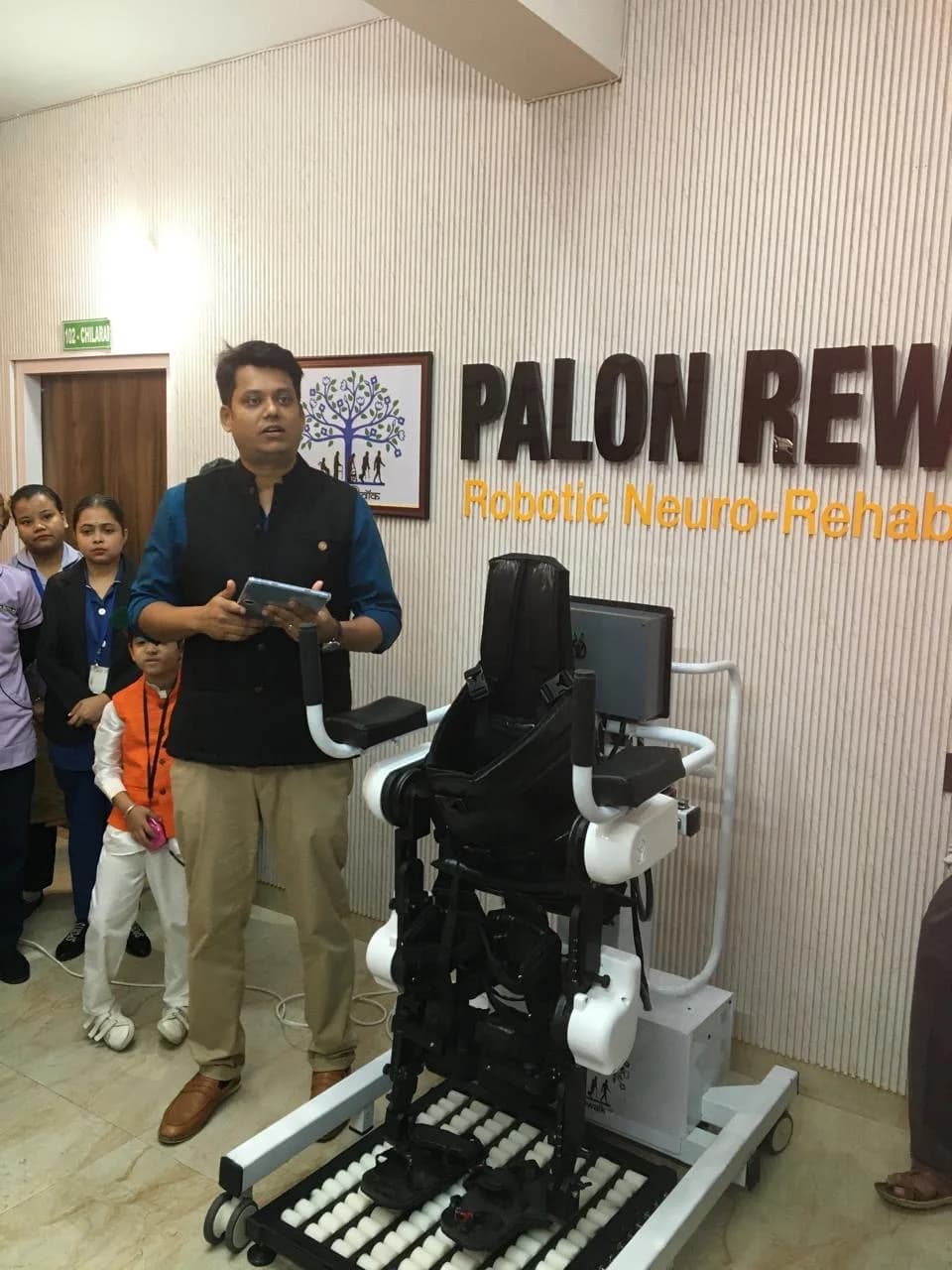A robotic gait-training exoskeleton mounted on a wheeled frame with a treadmill-like footplate, demonstrated by a staff member holding a tablet at a neuro-rehabilitation centre in Guwahati, India.