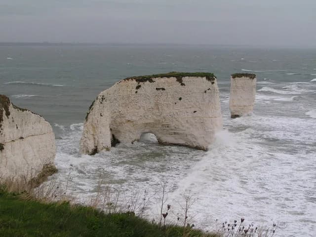 Photograph of chalk stacks at Old Harry Rocks, Dorset: a large stack with a small arch at its base and a slender stack beyond, separated from the chalk cliff of the headland by the sea.