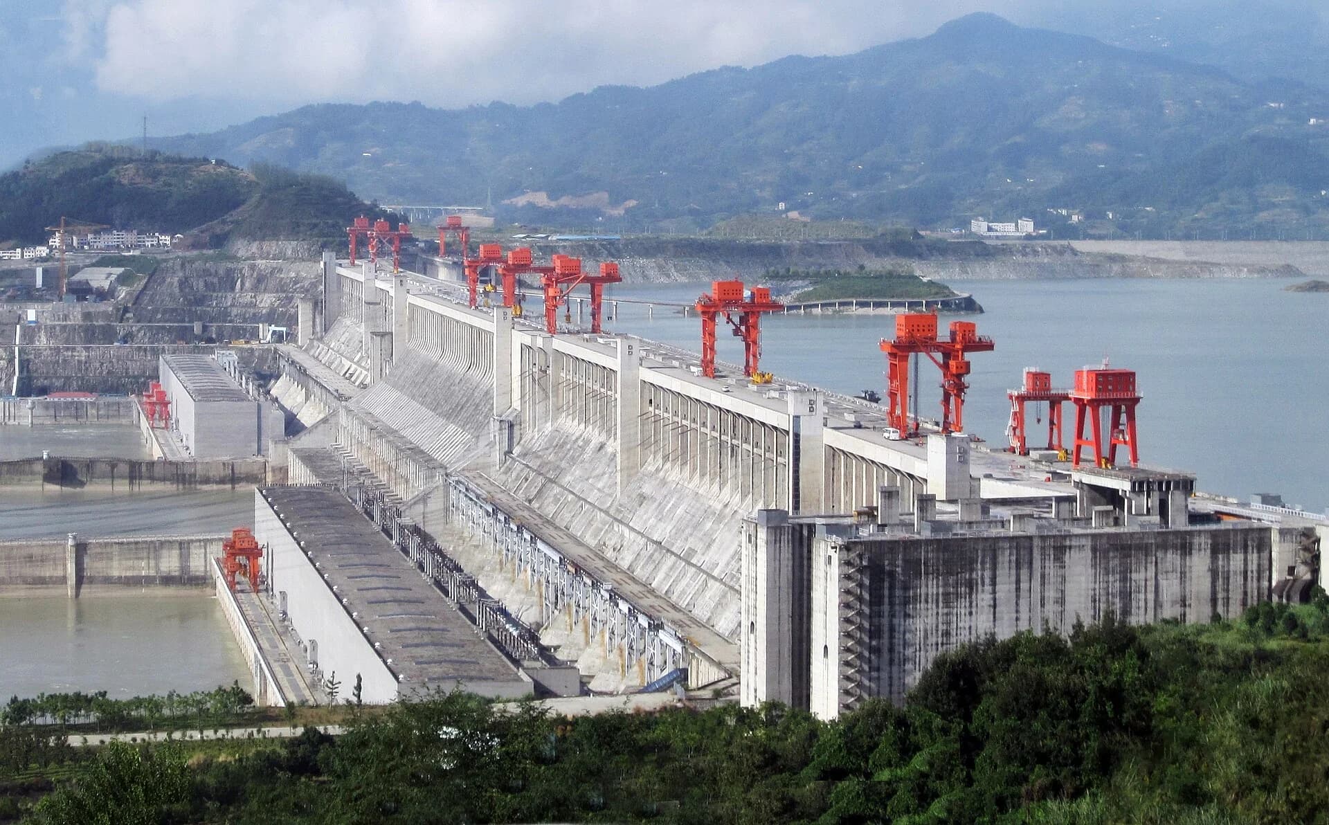 The Three Gorges Dam on the Yangtze River, China: a long concrete gravity dam with spillway gates and gantry cranes, the wide reservoir on the upstream side to the right and the lower river and power station on the downstream side to the left.