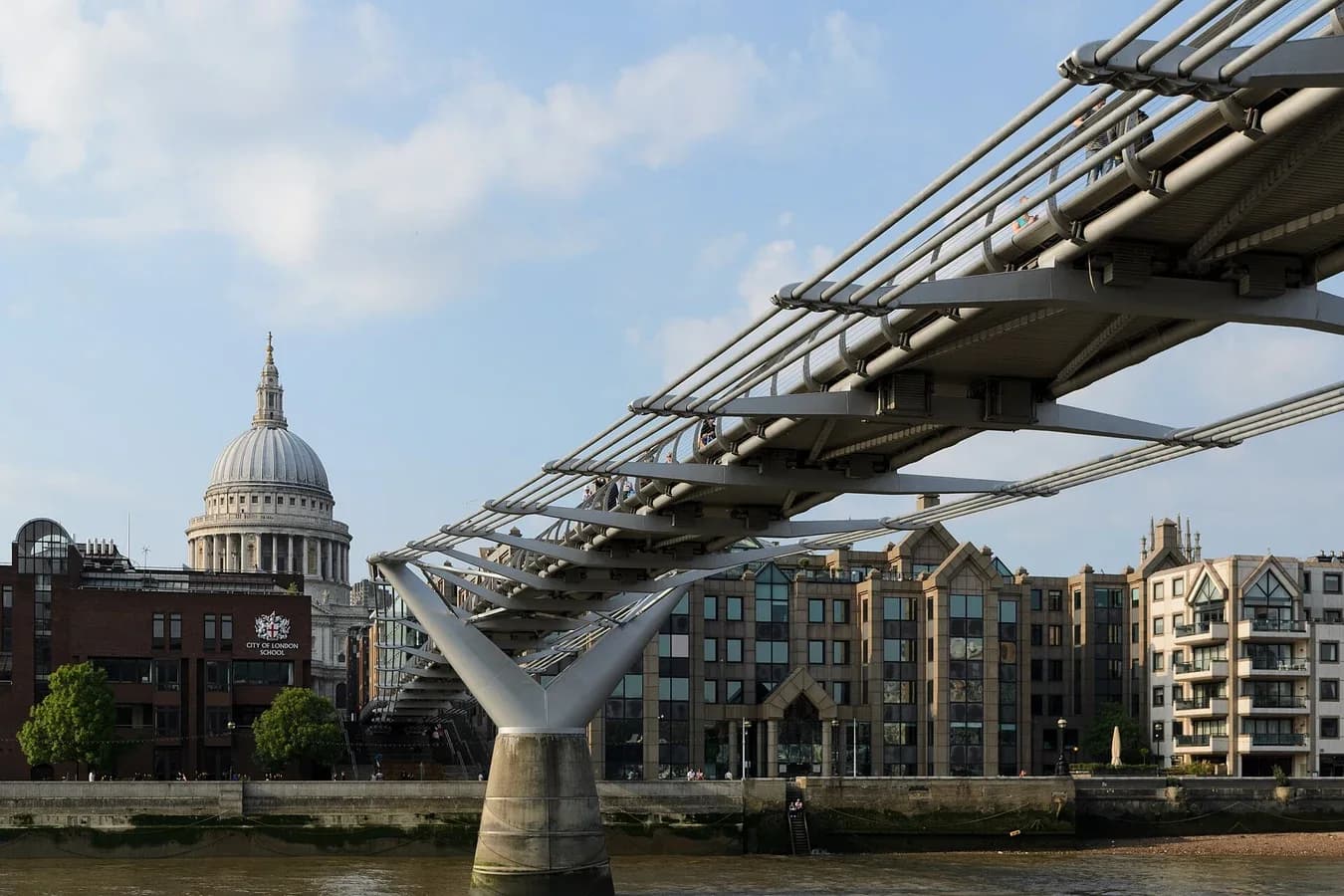 The Millennium Bridge in London seen from the river bank: a Y-shaped steel pier supports a slender deck, with bundles of shallow suspension cables running beside the deck rather than high above it. St Paul's Cathedral is in the background.