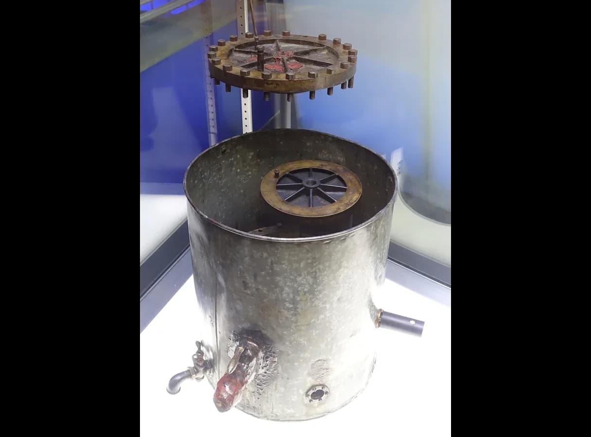 Photograph of Millikan’s original oil-drop apparatus in a museum case: a metal cylindrical chamber with its bolted lid raised above it, a round plate visible inside, and tubes and a tap on its side.