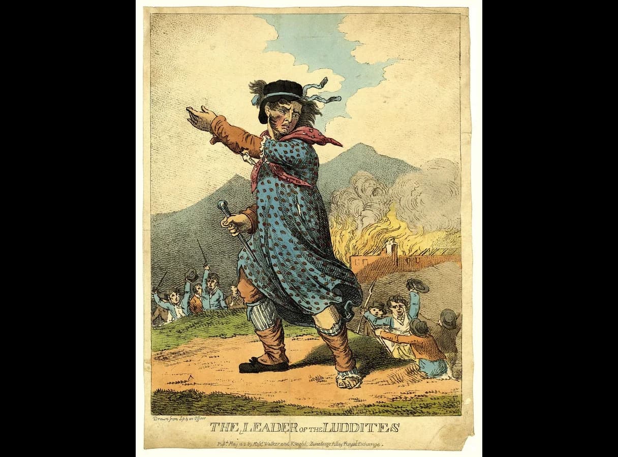 Hand-coloured print of a scowling man in a spotted woman's gown and shawl, with ribbons on his hat, pointing forward and holding a stick. Men waving hats and weapons follow him, and a building burns behind. The title reads The Leader of the Luddites.