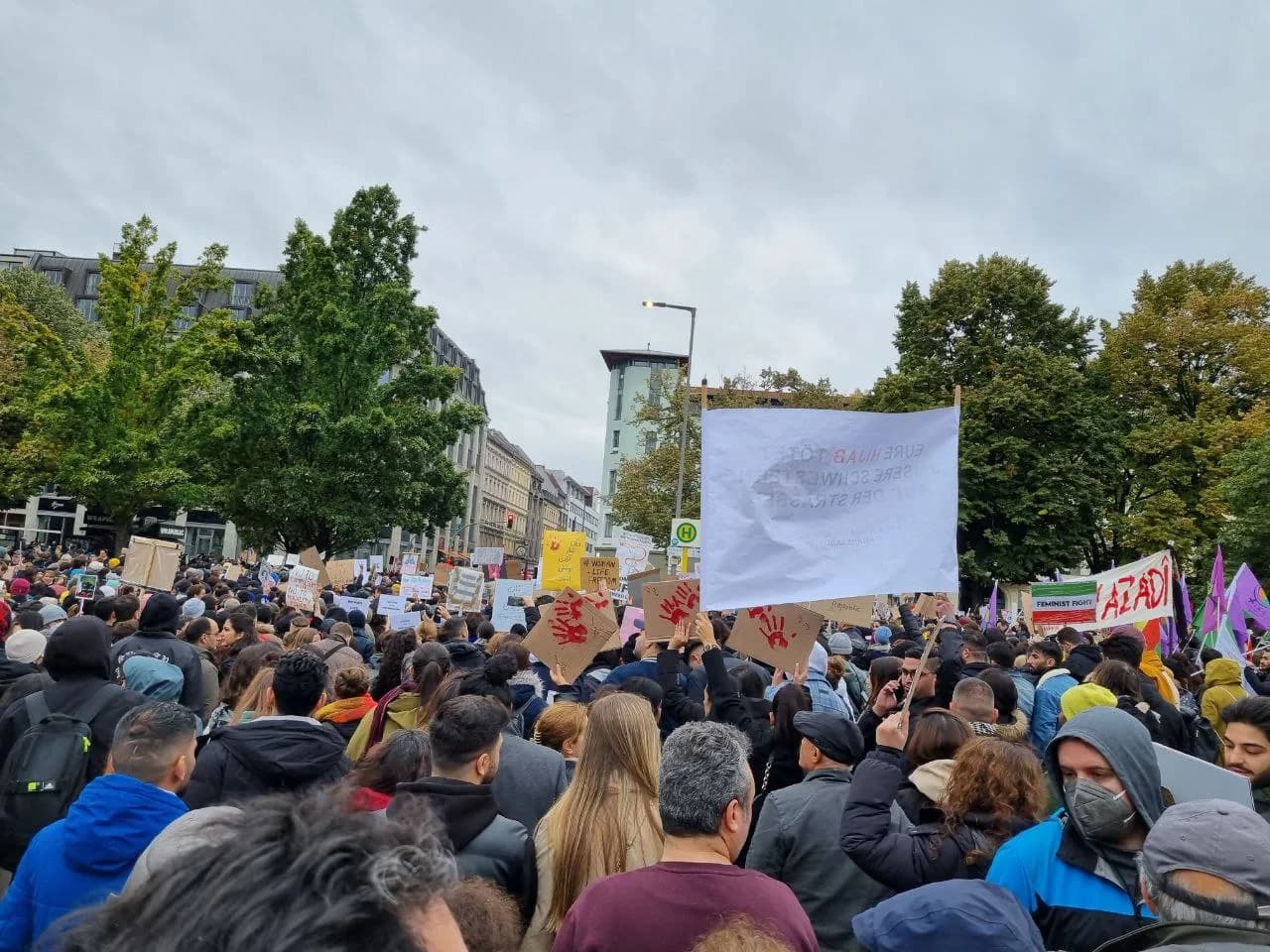A large crowd marching in a Berlin street in autumn 2022 in solidarity with the protests in Iran, holding placards with red handprints, a 'Woman Life Freedom' sign and a banner reading 'Azadi' (freedom).