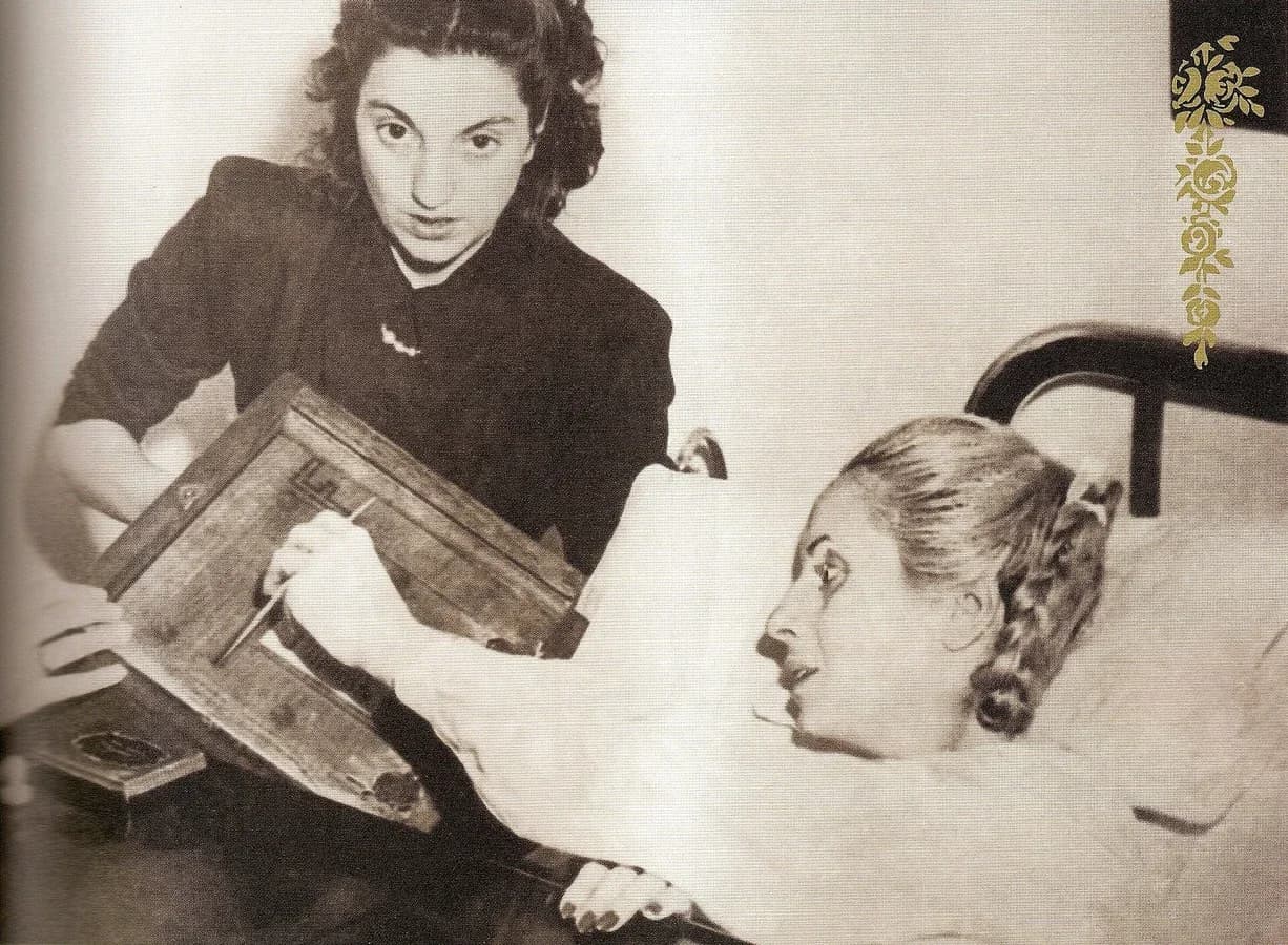 Sepia photograph of Eva Perón lying in a hospital bed, reaching out to place her ballot in a wooden ballot box held up beside the bed by a young woman in a dark dress.