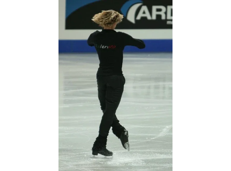 A figure skater spinning upright on one skate during practice at a championship, arms bent at shoulder height and free leg crossed in front, hair flung outwards by the rapid rotation.