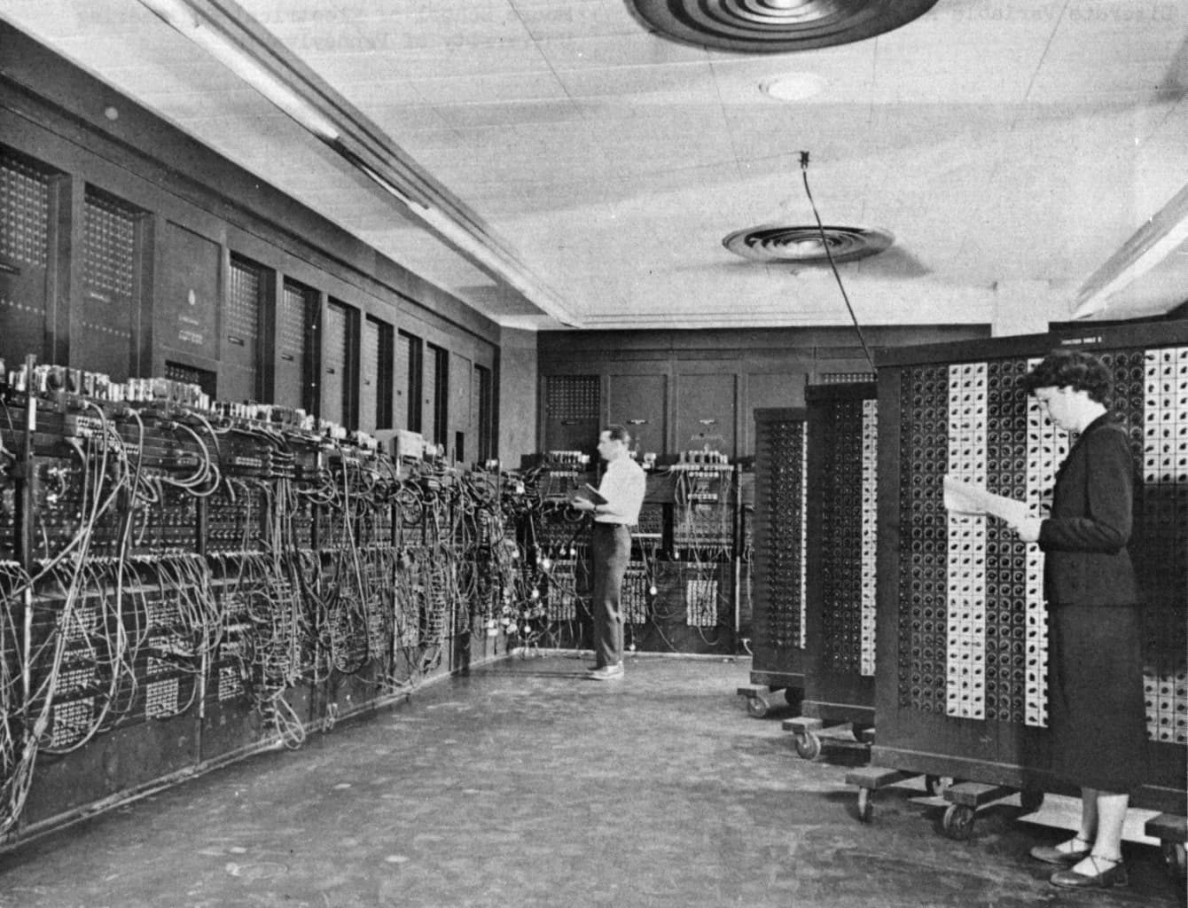 Black-and-white photograph of ENIAC: two programmers, a man in the background and a woman in the foreground, set switches and cables on tall panels of the room-sized computer.