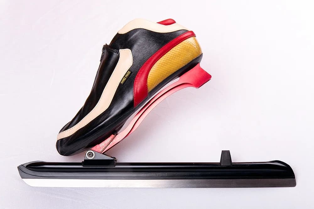 A clap skate seen from the side: a speed skating boot attached to a long blade by a hinge under the ball of the foot. The heel has lifted away from the blade, which stays flat on the ground.
