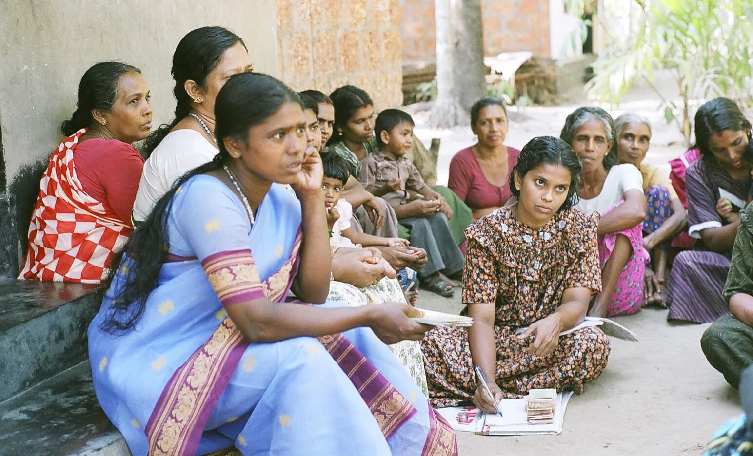 A women's self-help microfinance group meeting in Kerala, India, with members sitting together and recording savings and loans in ledgers.