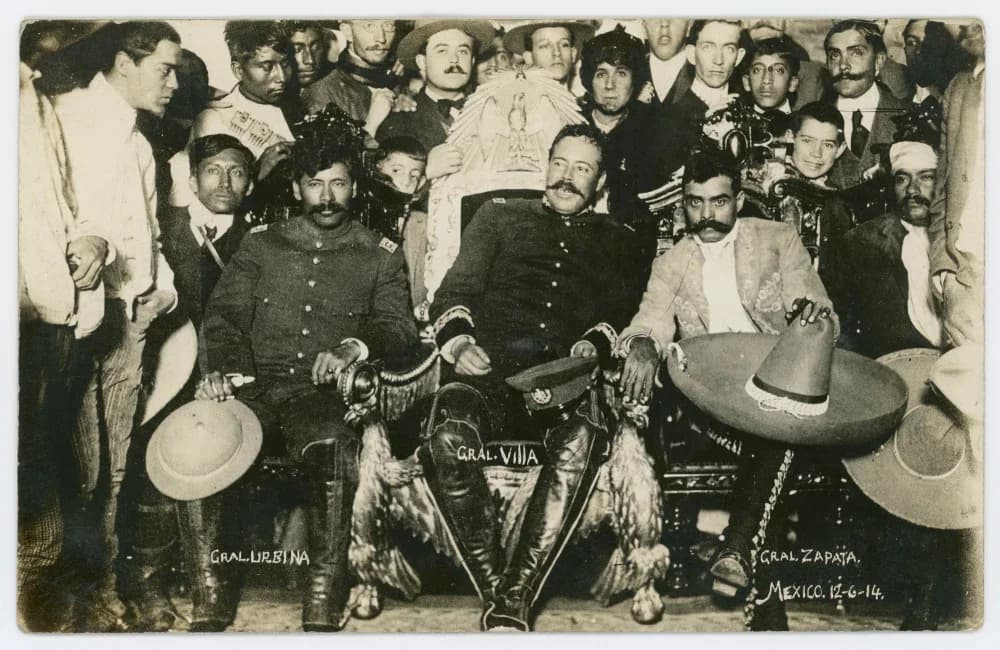 Photograph of Pancho Villa sitting in the ornate presidential chair in the National Palace, with General Tomás Urbina on his left and Emiliano Zapata, holding a large sombrero, on his right, surrounded by a crowd of men and boys.
