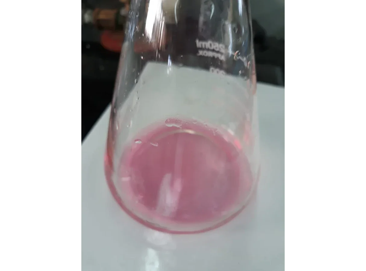Photograph of a conical flask standing on a white surface. The solution in it is a faint pink colour, the end point of a titration with phenolphthalein indicator.