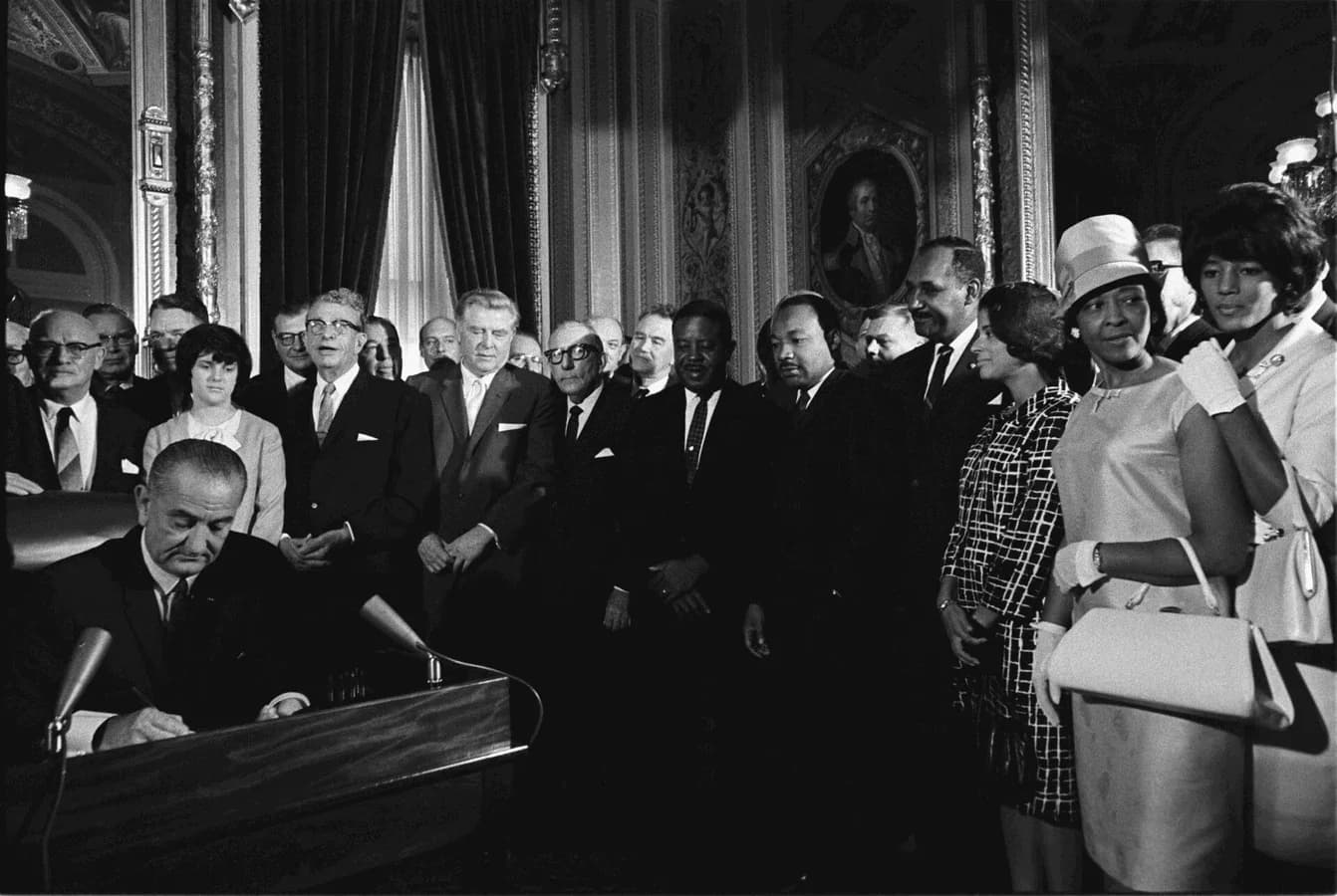 Black-and-white photograph of President Lyndon Johnson seated at a desk signing a document, surrounded by standing men and women, including Martin Luther King Jr.