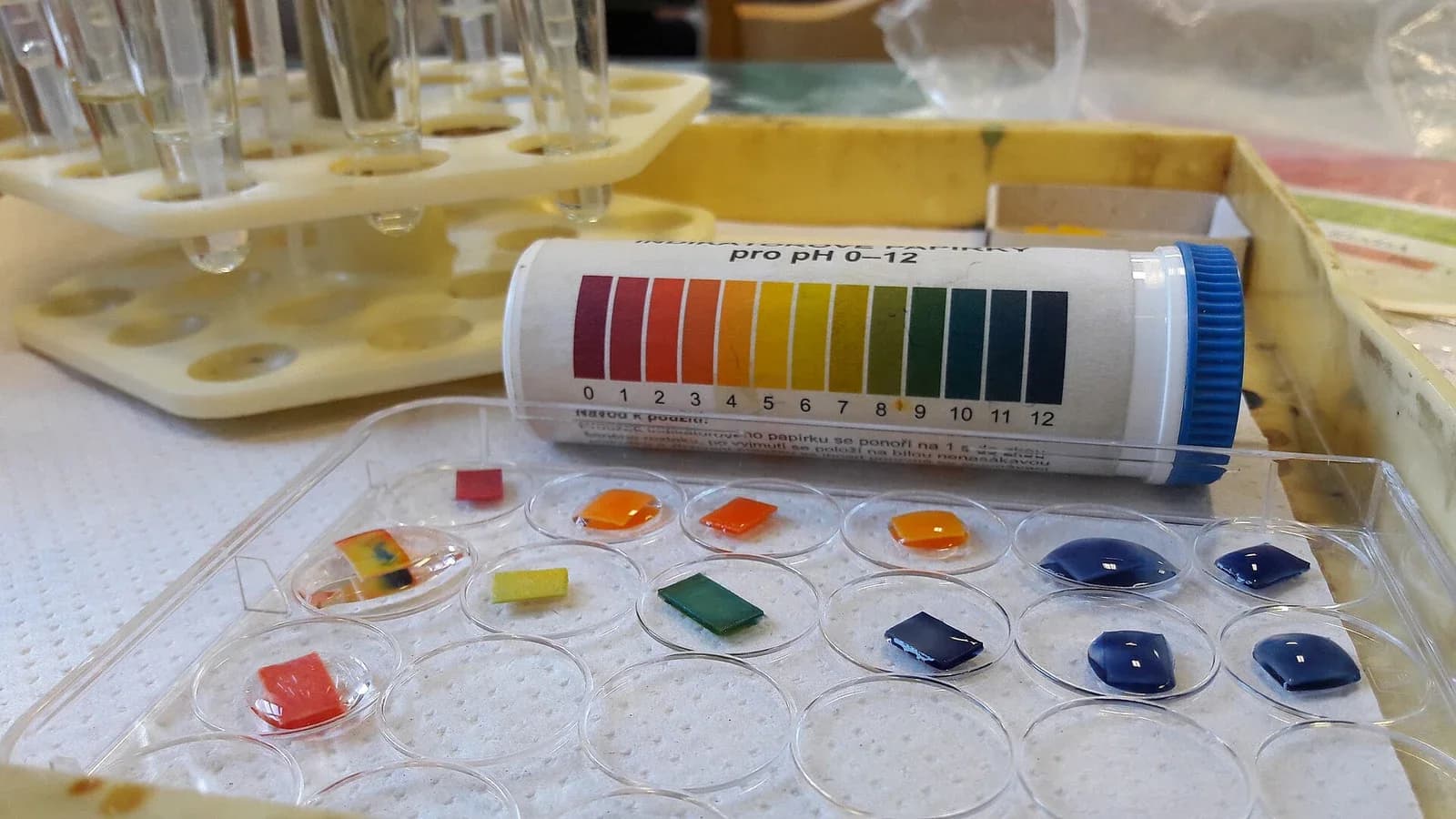 A tube of universal indicator paper with its printed colour chart for pH 0 to 12 (red through orange, yellow and green to dark blue), and small squares of the paper on a dish, each dipped in a different solution and showing red, orange, yellow, green or blue.