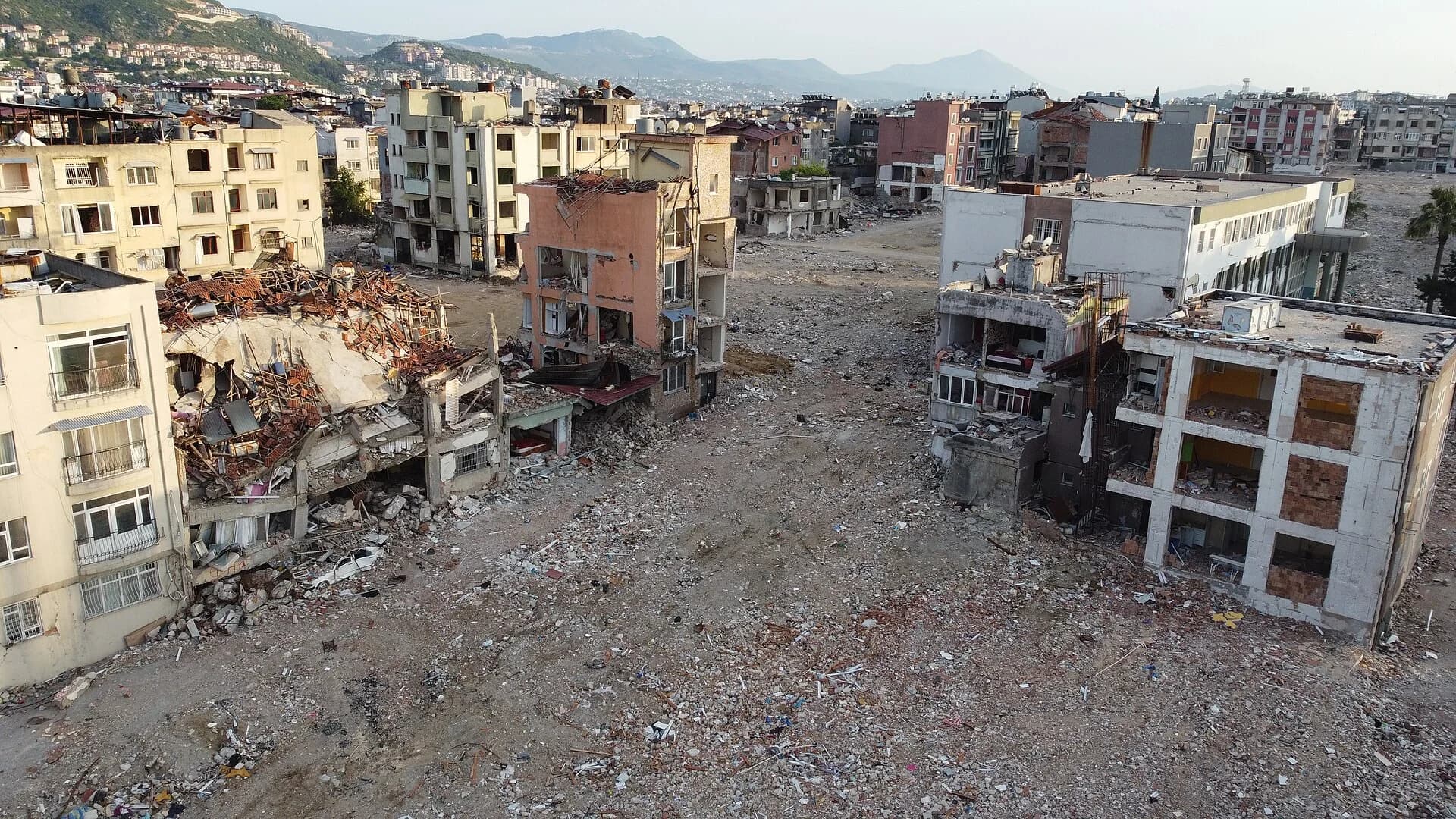 Aerial photo of central Antakya after the February 2023 earthquakes: collapsed and partly collapsed apartment blocks beside cleared plots of rubble, with standing damaged buildings and hills behind.