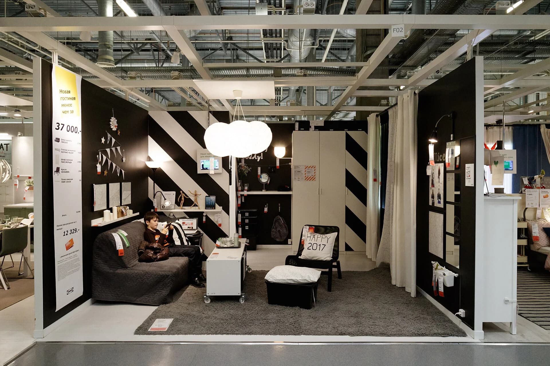 A furnished living-room set inside an IKEA store in Rostov-on-Don, Russia, with sofa bed, shelves, lamps and wardrobe, price tags on each item and a large board giving the total price of the whole room.