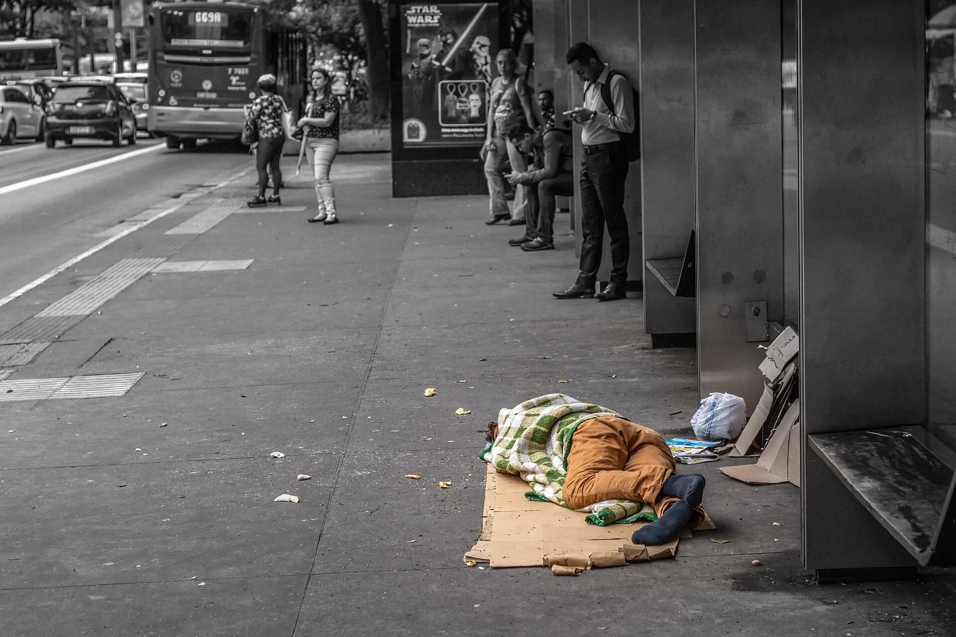 A person asleep on flattened cardboard under a blanket on a São Paulo pavement, while office workers in shirts wait at a bus stop a few metres away.