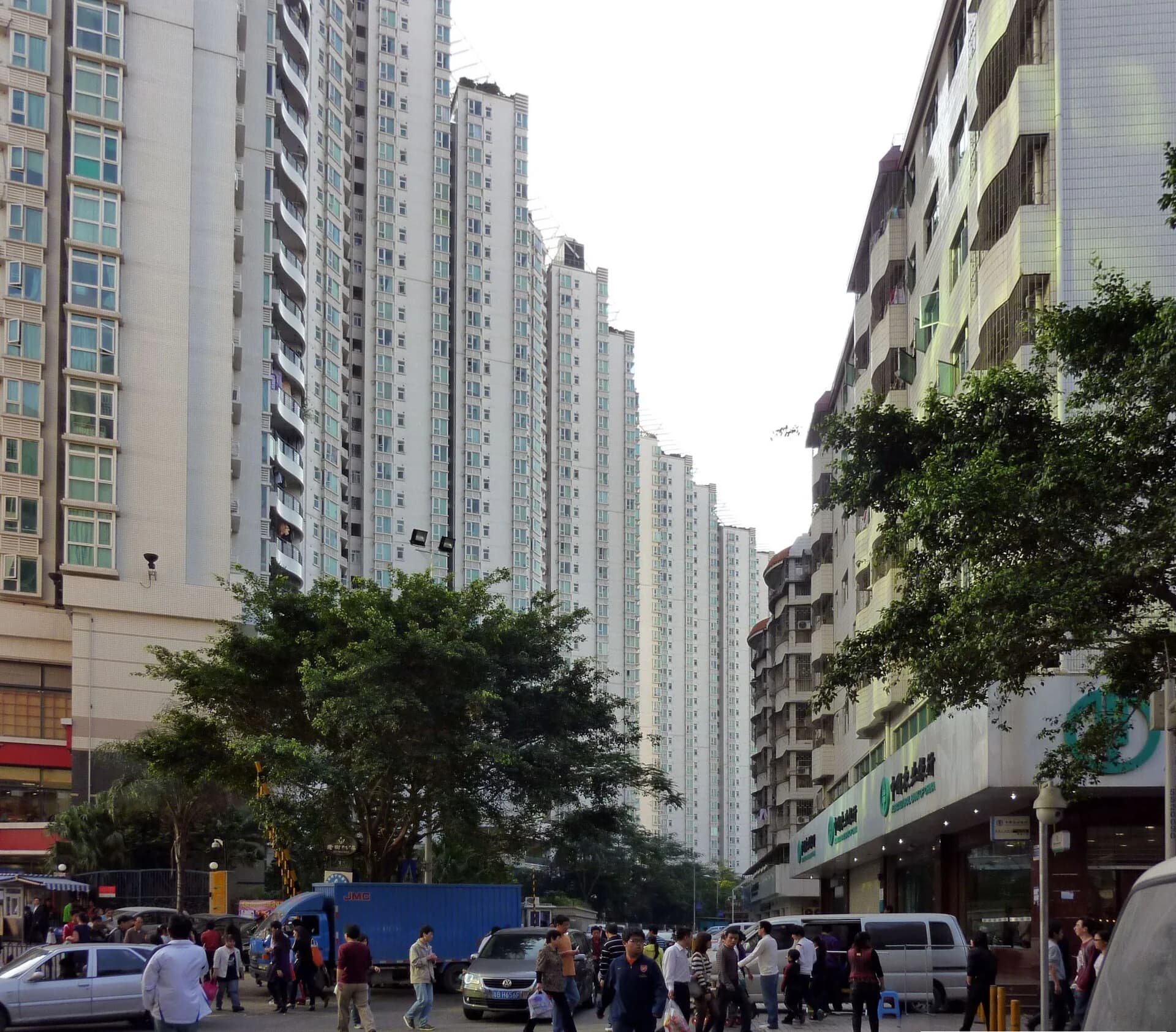 Street in Baishizhou, Shenzhen: older six- to eight-storey village buildings with barred balconies on the right face a row of 30-storey modern apartment towers, with crowds and vans on the street.