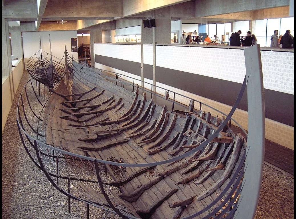 The surviving timbers of a Viking cargo ship displayed in a museum hall. Dark overlapping planks form a broad, deep hull with heavy cross-beams and ribs; a metal frame outlines the missing parts of the original shape.