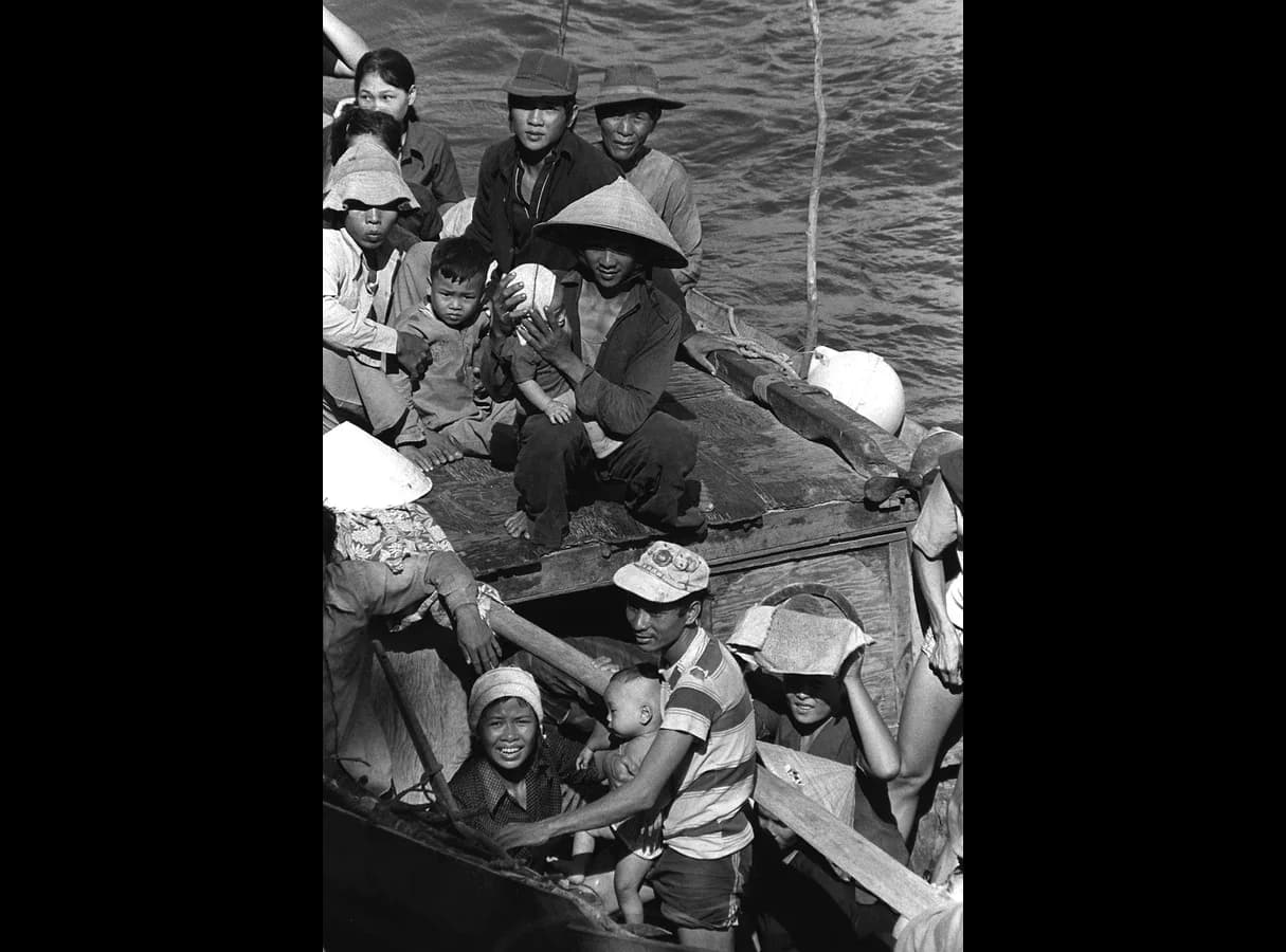 Black-and-white photograph taken from above: a small wooden fishing boat at sea, crowded with men, women and young children sitting on the deck and standing in the open hold, some in conical hats.