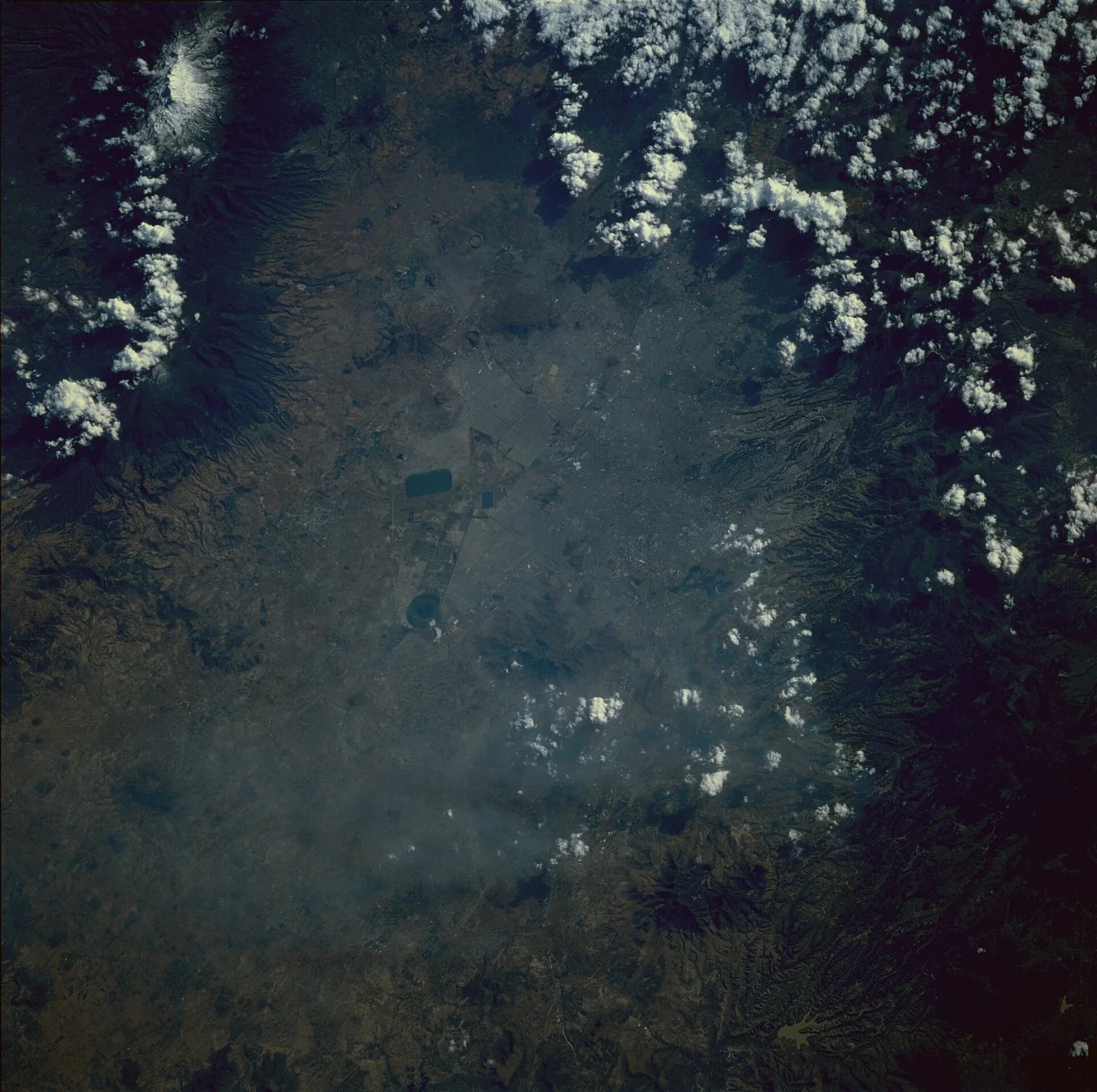 Space Shuttle photograph of the Valley of Mexico in November 1985: a grey-brown haze of smog covers the city on the basin floor, surrounded by darker forested mountains and volcanoes.