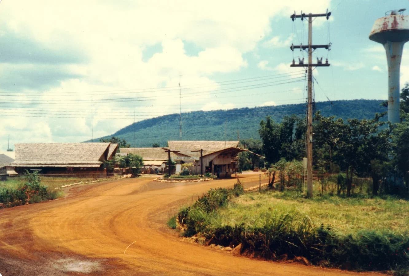 Colour photograph of a red dirt road curving to the thatched buildings and gatehouse at the entrance of Khao-I-Dang camp, with a forested mountain behind, power lines and a water tower.