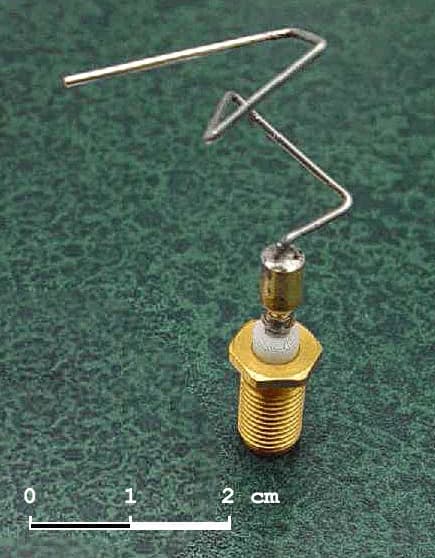 Photograph of a small bent-wire antenna with several irregular kinks, mounted on a gold-coloured threaded connector, next to a 2 cm scale bar.