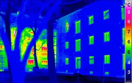 Infrared thermal image on a colour scale from 3 °C (blue) to 9 °C (pink). On the right, a well-insulated passive house has blue walls with slightly warmer green windows. On the left, behind trees, an older building shows green, yellow and red walls and windows.