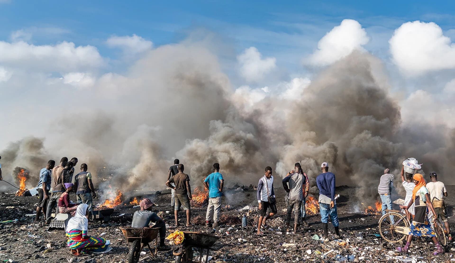 Photograph of the Agbogbloshie scrap site in Accra, Ghana, in 2019: people stand among small fires and thick grey smoke where cables and plastic are burned on blackened ground, with wheelbarrows and a bicycle in the foreground.