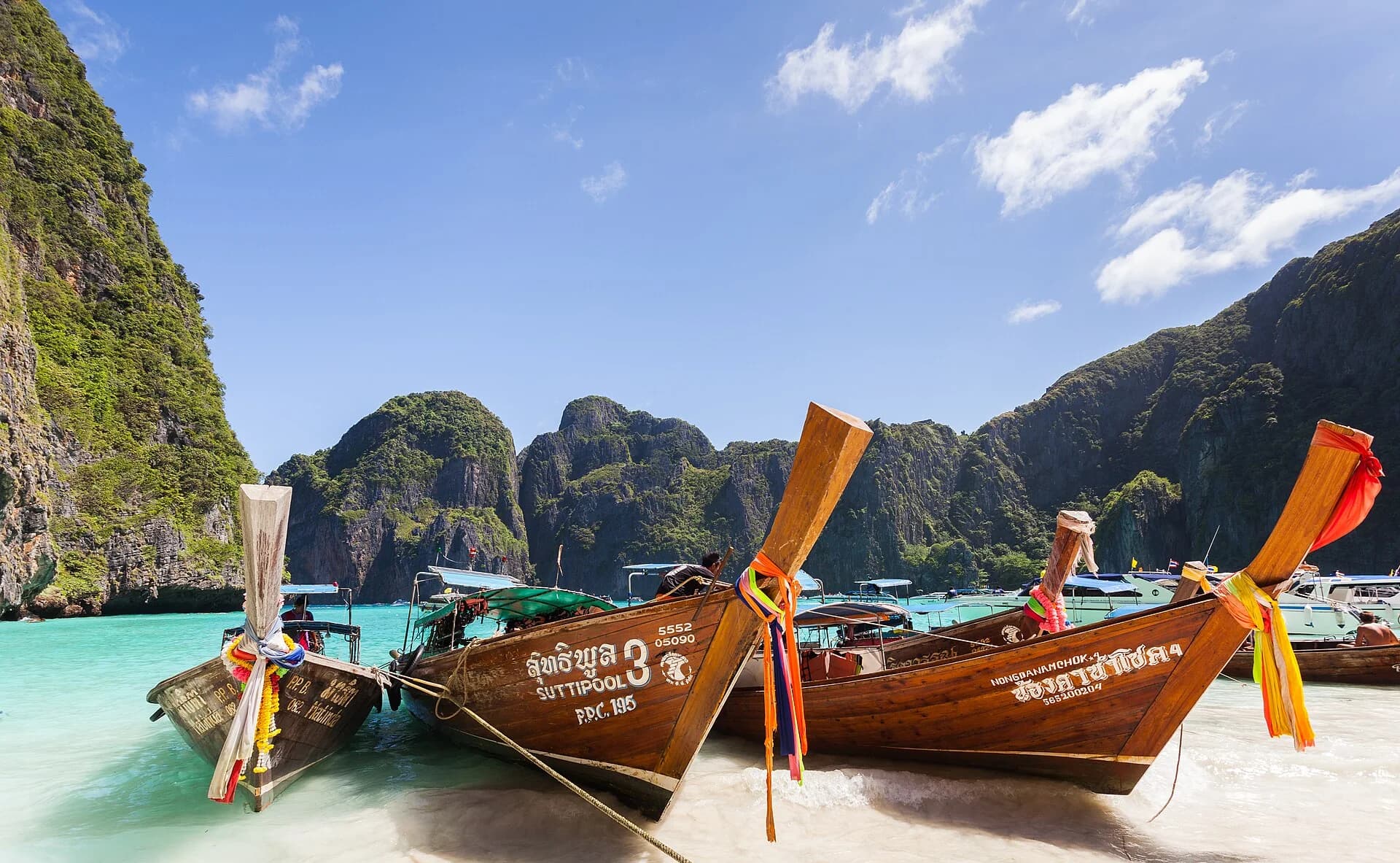 Long-tail boats moored side by side on the beach at Maya Bay, Ko Phi Phi Le, Thailand, in 2013, with more boats and tourists behind and limestone cliffs around the bay.