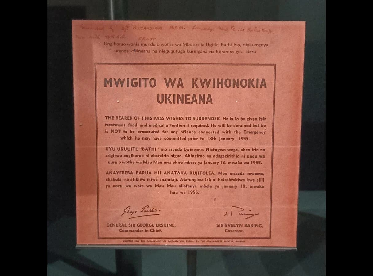 A printed red surrender pass of 1955 in Kikuyu, English and Swahili, signed by General Sir George Erskine and Governor Sir Evelyn Baring. The English text says the bearer wishes to surrender, is to be given fair treatment, food and medical care, and will be detained but not prosecuted for…