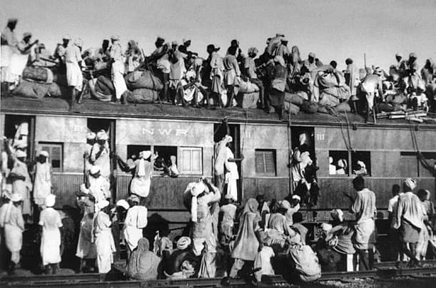 Black-and-white photograph of a train so crowded with refugees that people sit packed on the carriage roofs and hang from the sides.