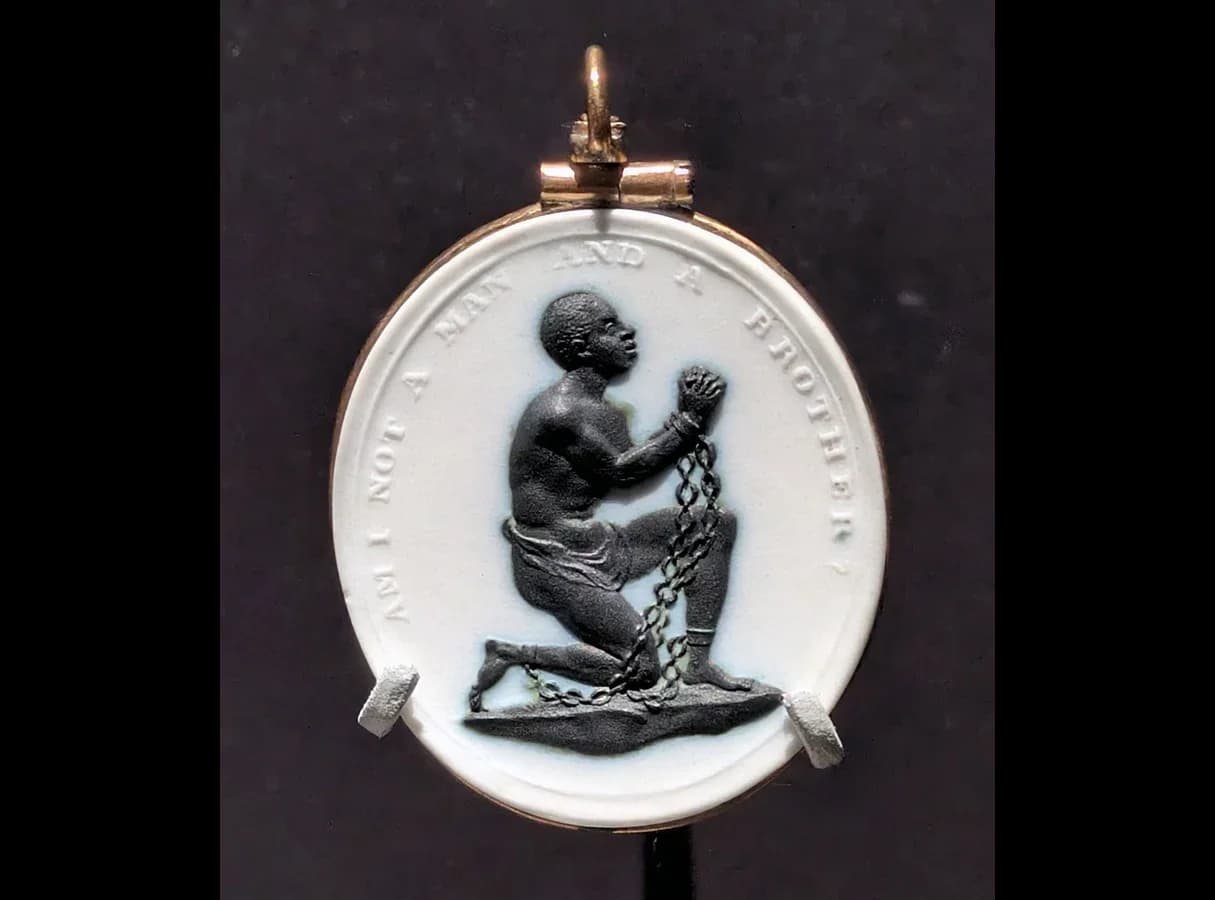 Oval jasperware cameo in a gold mount: a black relief of a kneeling African man in chains, raising his clasped hands, on a white ground; around the edge runs the motto "Am I not a man and a brother?".