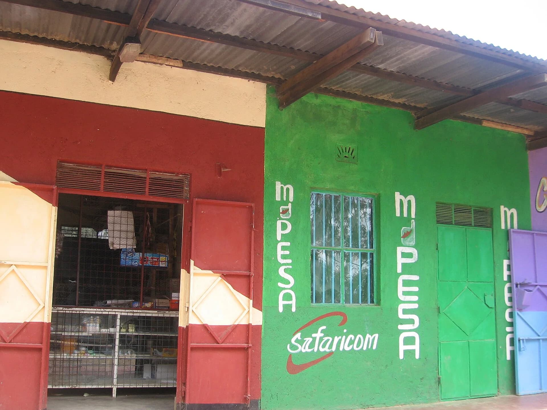 A roadside shop in Kenya painted green with M-PESA and Safaricom logos, next to a small general store with a red door.